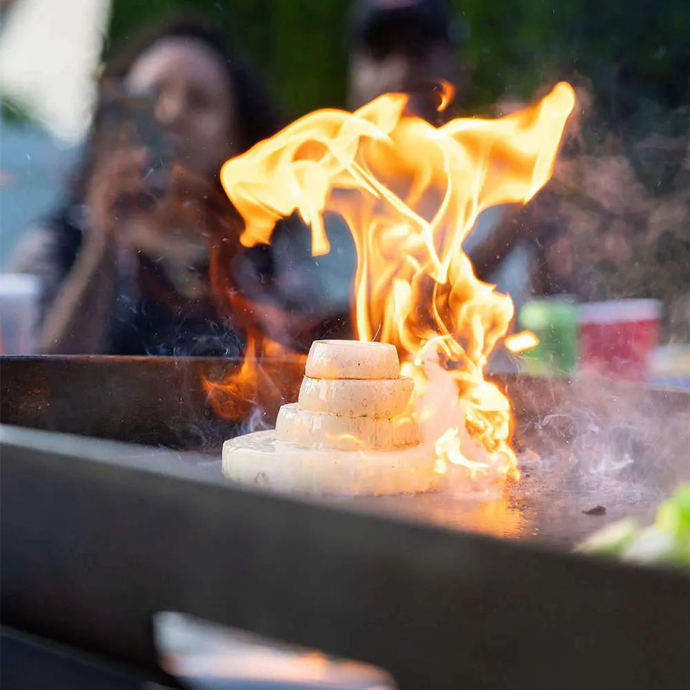 Flaming onion volcano trick on an outdoor flat-top grill with people watching in the background.