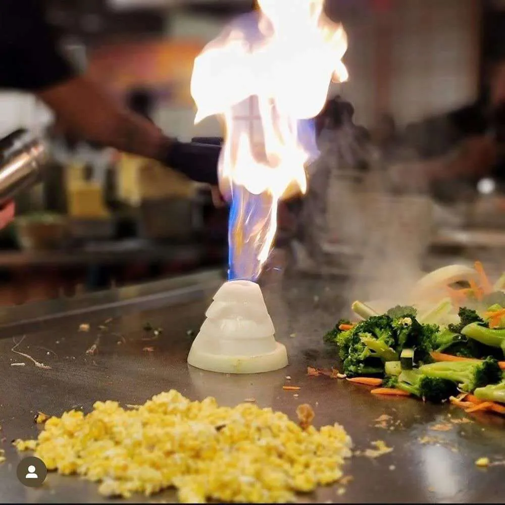 Hibachi chef performing a flaming onion volcano trick on a griddle with fried rice and vegetables.