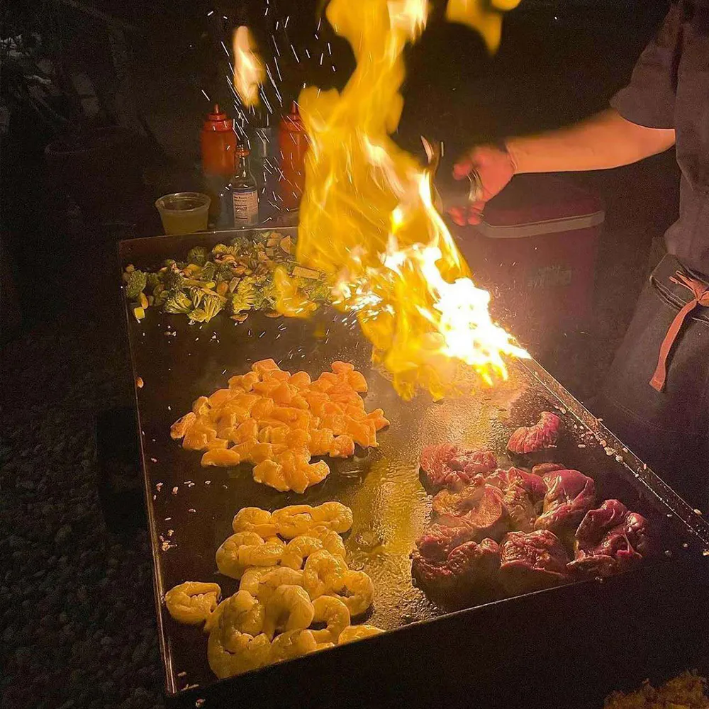 Huge flames erupting from an outdoor flat-top grill cooking shrimp, steak, and vegetables.
