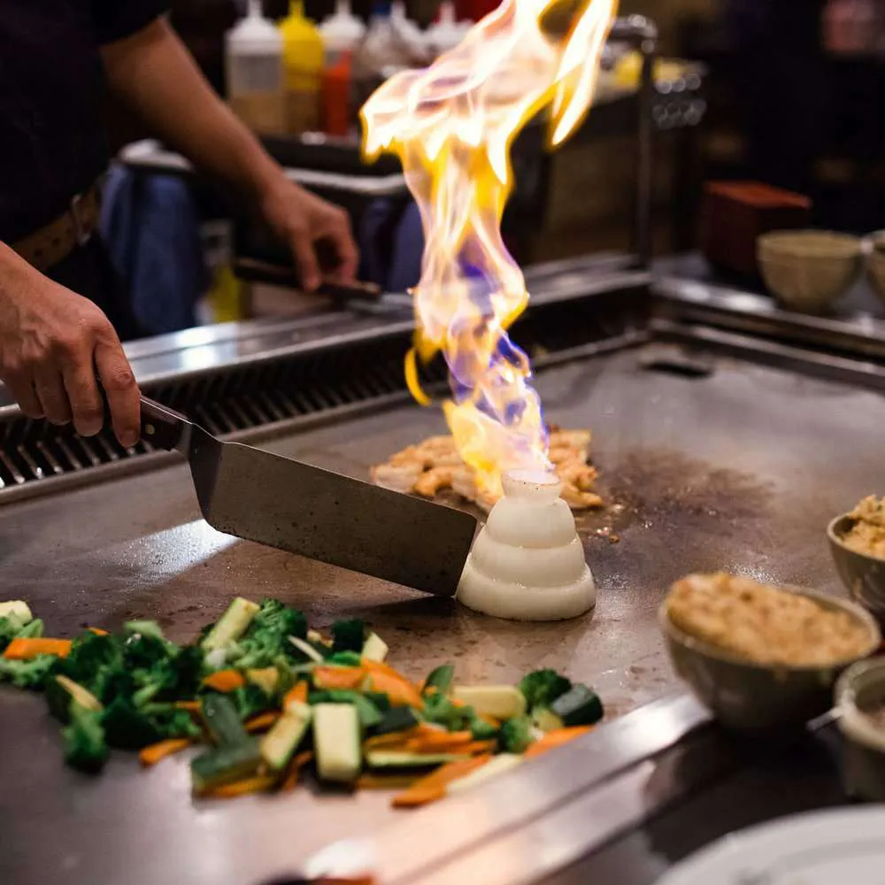 Teppanyaki chef creating a large fire from an onion volcano on a flat-top grill with mixed vegetables.